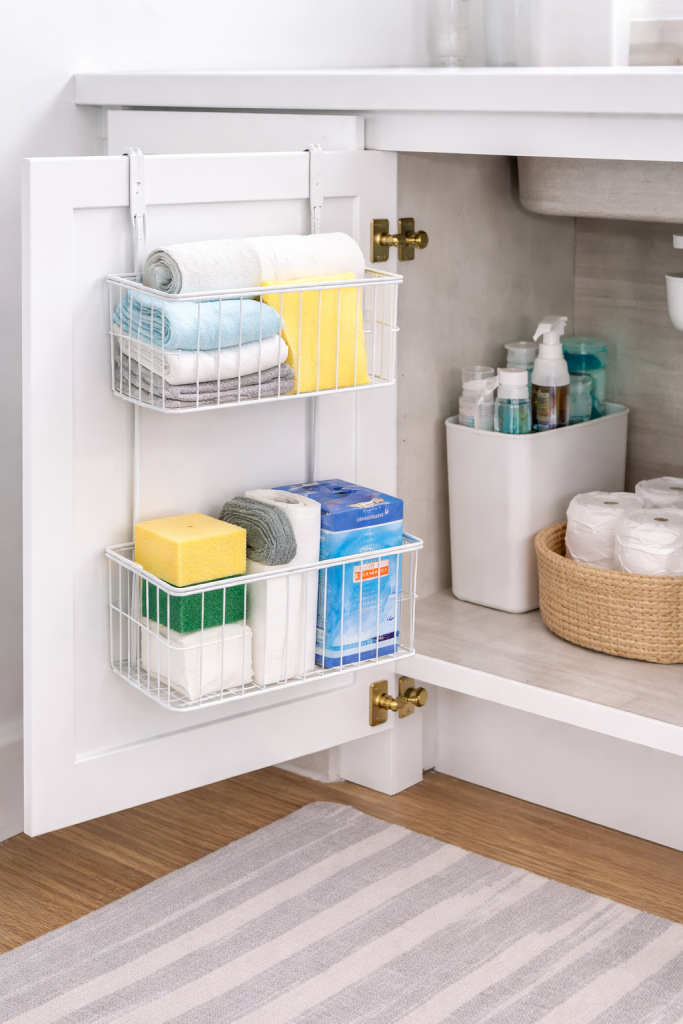 Hanging basket organizer on cabinet door storing kitchen cloths sponges and garbage bags to reduce clutter under the sink